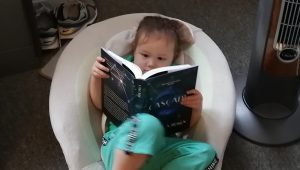 Photo of small child reclining in a bouncy chair while holding hard-cover copy of Rachel A. Rosen's novel, Cascade.