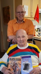 Image shows Geoffrey Dow, standing behind Carl Dow, who is holding copies of his books, Black Grass, and The Old Man's Last Sauna, while seated in his wheelchair. Photo by Judy Kwasnica.