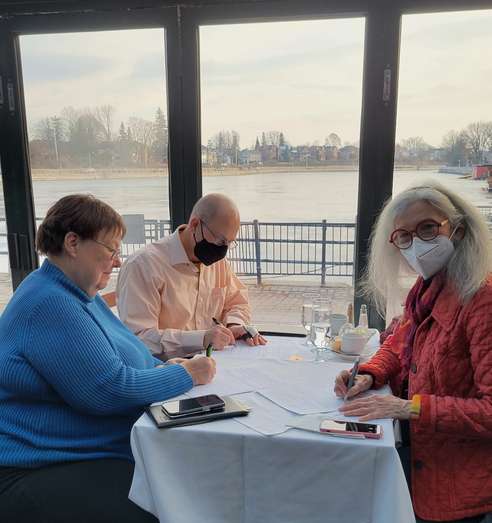 Image is a photo of Marie-Andrée Auclair, Geoffrey Dow, and Adrienne Stevenson, each signing a contract with the Rideau Canal appearing in the background.