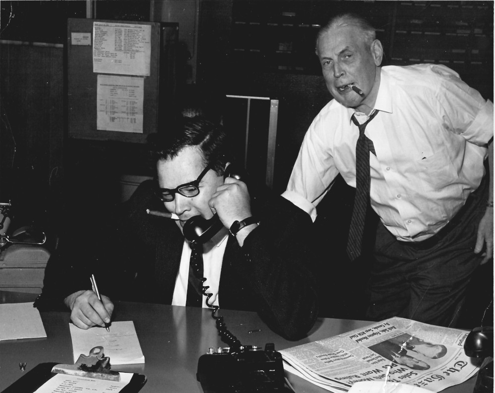 Photo shows Carl Dow taking notes while on the phone, as an older man looks over his shoulder. Both are smoking cigarillos.